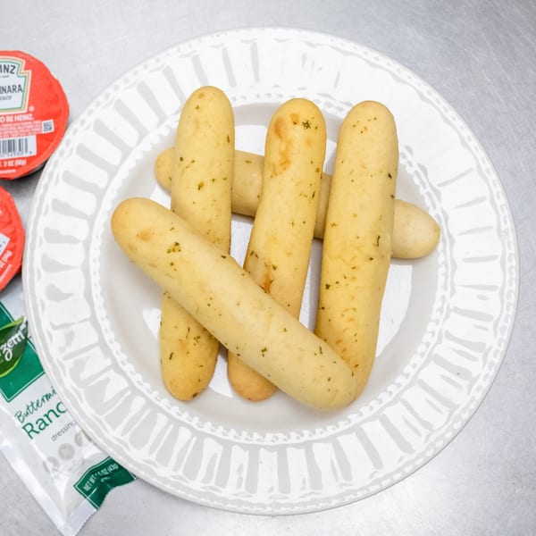 A white plate holds several golden-brown breadsticks, with small containers of Heinz marinara and ranch dressing nearby.