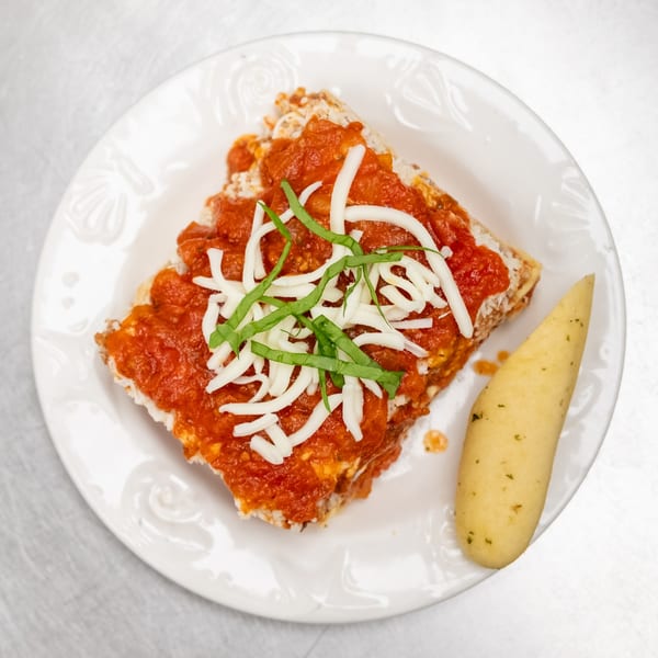 A slice of lasagna topped with red sauce, shredded cheese, and basil, with a breadstick.