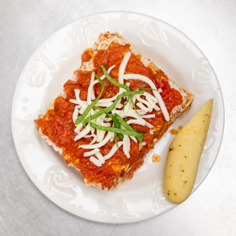 A slice of lasagna topped with red sauce, shredded cheese, and basil, with a breadstick.
